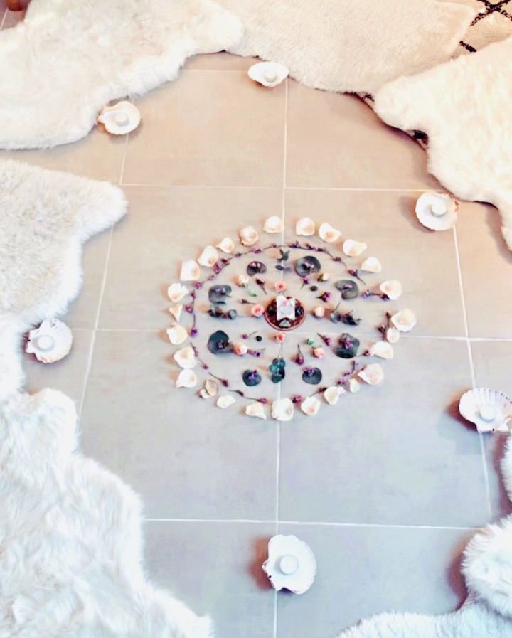 cercle sur le sol fait avec des pétales de fleurs de couleurs naturelles, coqulles St Jacques contenant des bougies et des fausses peaux de bêtes en fourrure pour s'assoir.