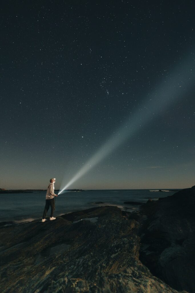 Homme portant une casquette et marchant sur des rochers en bord de mer de nuit. Il tient une lampe torche et regarde vers le haut.