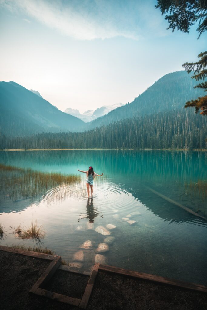 image d'un lac sauvage entouré d'arbre, avec une jeune femme brune marchant dans l'eau avec les bras écartés.